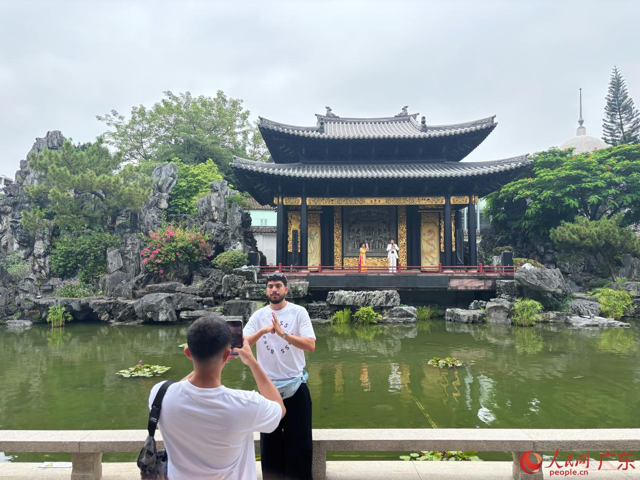 临水戏台粤剧正演，外国游客站在池边，以粤剧表演为背景拍照留念。人民网 李舜摄